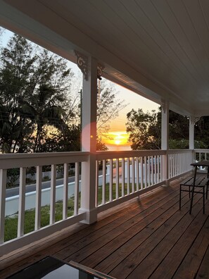 A wooden deck with a railing, overlooking a road and trees, with a sunset in the background.