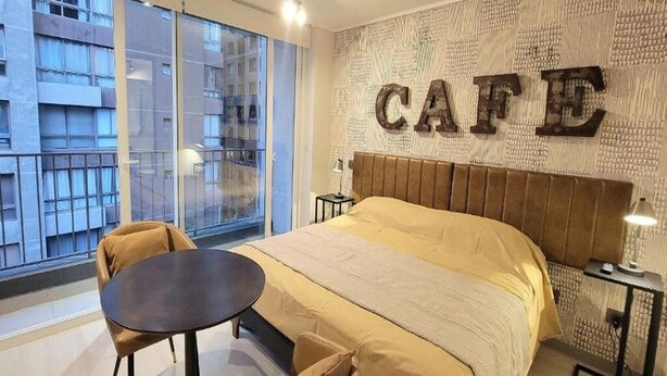 Ein modernes Hotelzimmer mit einem großen Bett, einem kleinen Tisch und Blick auf eine Stadtkulisse durch raumhohe Fenster.