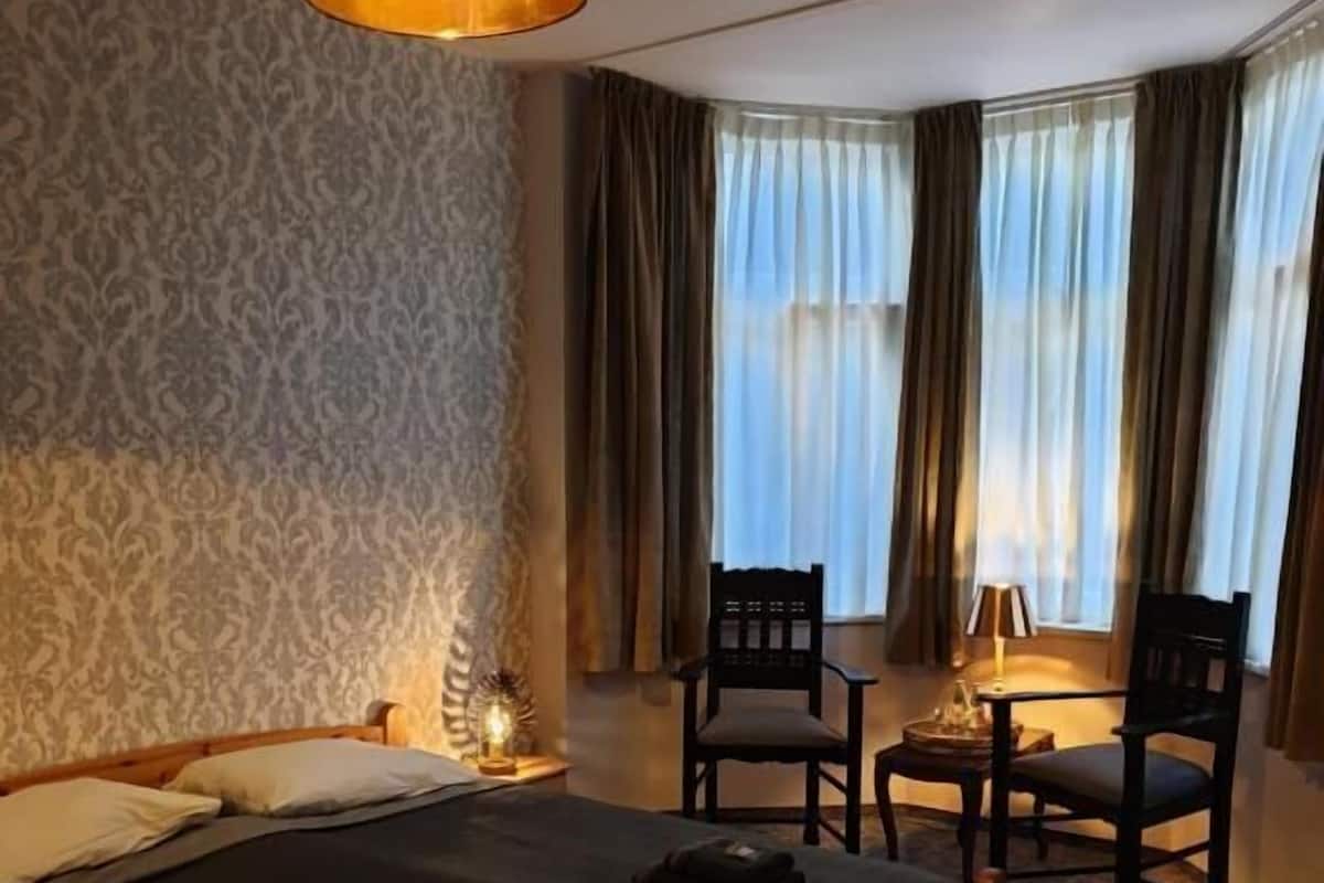 Une chambre d’hôtel avec un lit, deux chaises, une lampe et une fenêtre avec des rideaux.