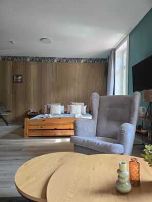 Una habitación con una cama, un sillón gris, una mesa redonda de madera y un ventanal con cortinas.