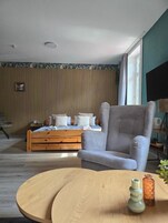 Una habitación con una cama, un sillón gris, una mesa redonda de madera y un ventanal con cortinas.