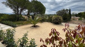 Ein angelegter Garten mit verschiedenen Bäumen und Sträuchern, einem Sandbereich und Blick auf entfernte Gebäude.