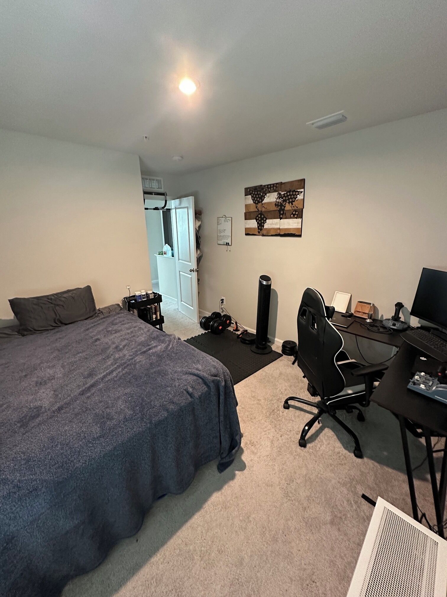 A bedroom with a bed, a desk with a computer, a chair, and a door leading to another room.