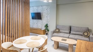 A modern living room with a sofa, coffee table, and chairs, a TV mounted on a patterned wall, and a wooden partition. - Ernest Suites Monastiraki (Athina)