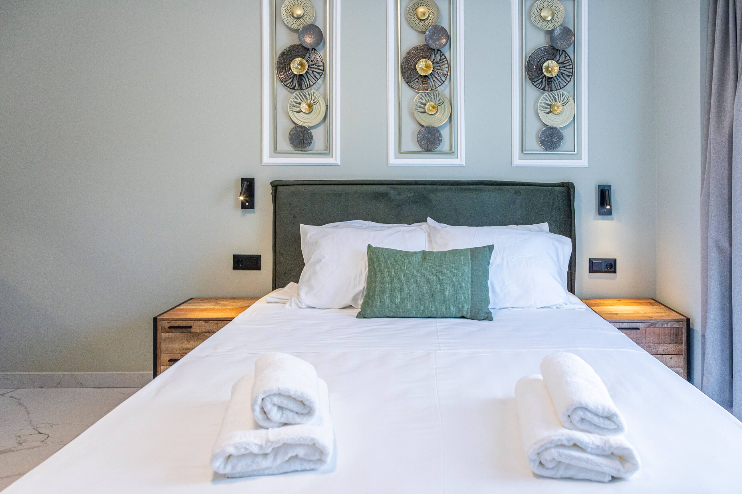 Een net opgemaakt bed met witte lakens en twee opgerolde handdoeken, een groen sierkussen en decoratieve wandkunst erboven.