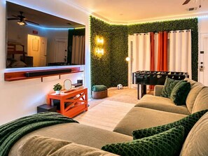 A living room with a sofa, a coffee table, a mirror, a wall-mounted shelf, a ceiling fan, and a wall with green foliage.