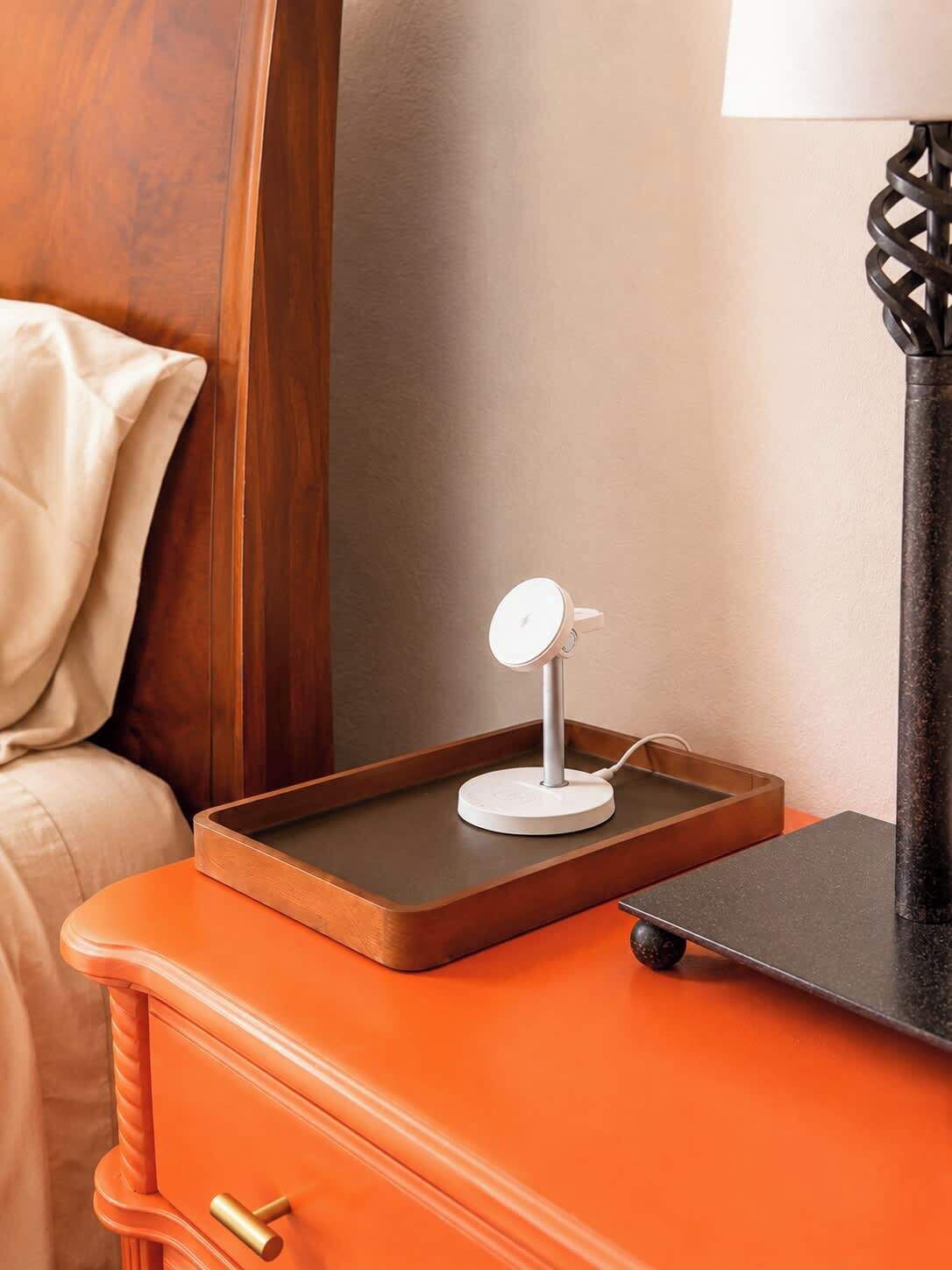 Una cama bien tendida con cabecera de madera, una mesita de noche con una lámpara, y una pequeña bandeja con un objeto blanco encima.