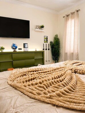 A bedroom with a large bed, a flat-screen TV mounted on the wall, a globe, a plant, and a bedside table with a lamp.