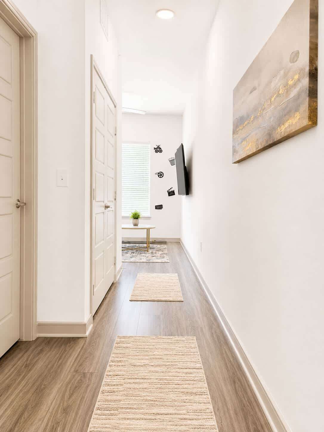 A hallway with a wooden floor, white walls, a door, a small table with a plant, and a mounted TV.