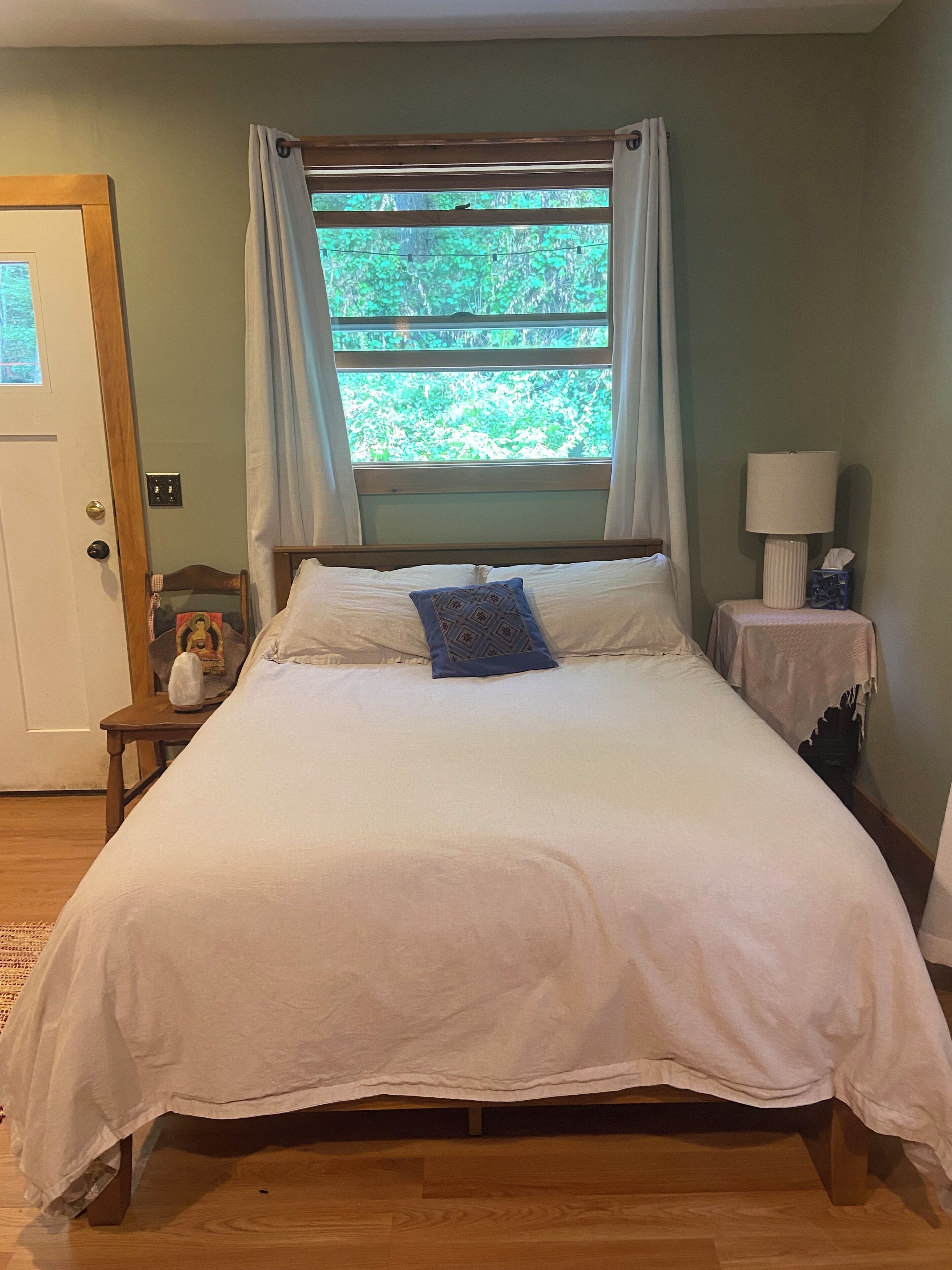 A bedroom with a bed, a nightstand with a lamp, a window with curtains, and a door.