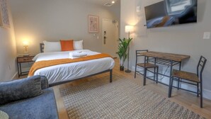 Un dormitorio con una cama, mesitas de noche, un escritorio con sillas, un televisor y una planta.