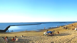 Ein Strand mit Menschen, einer sich ins Wasser erstreckenden Mole und einem wolkenlosen Himmel.