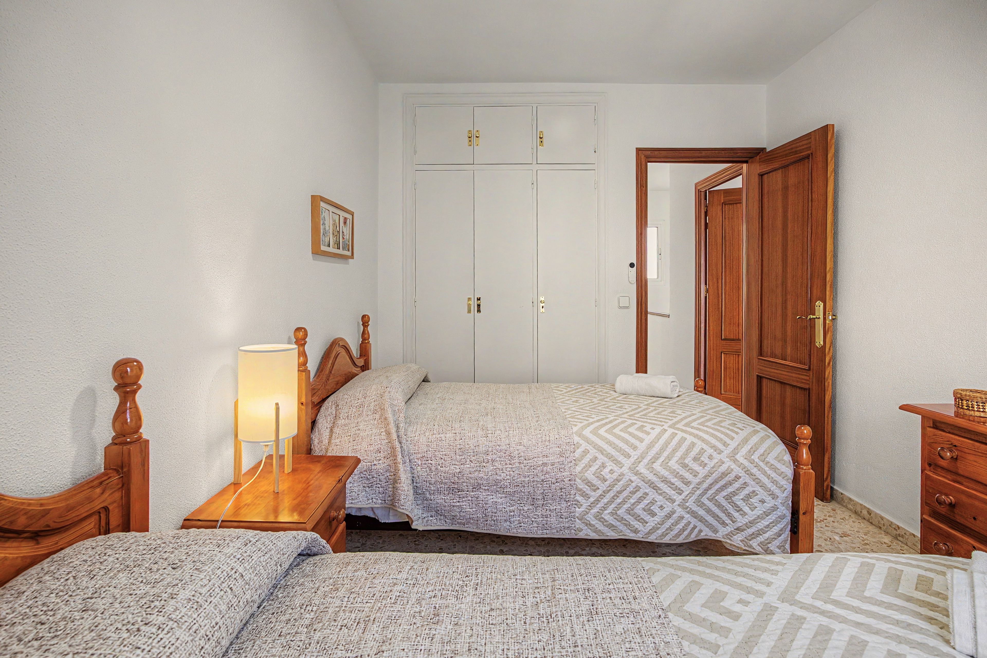 A bedroom with two single beds, a wooden nightstand, a bedside table with a lamp, a wardrobe, and a wooden dresser.