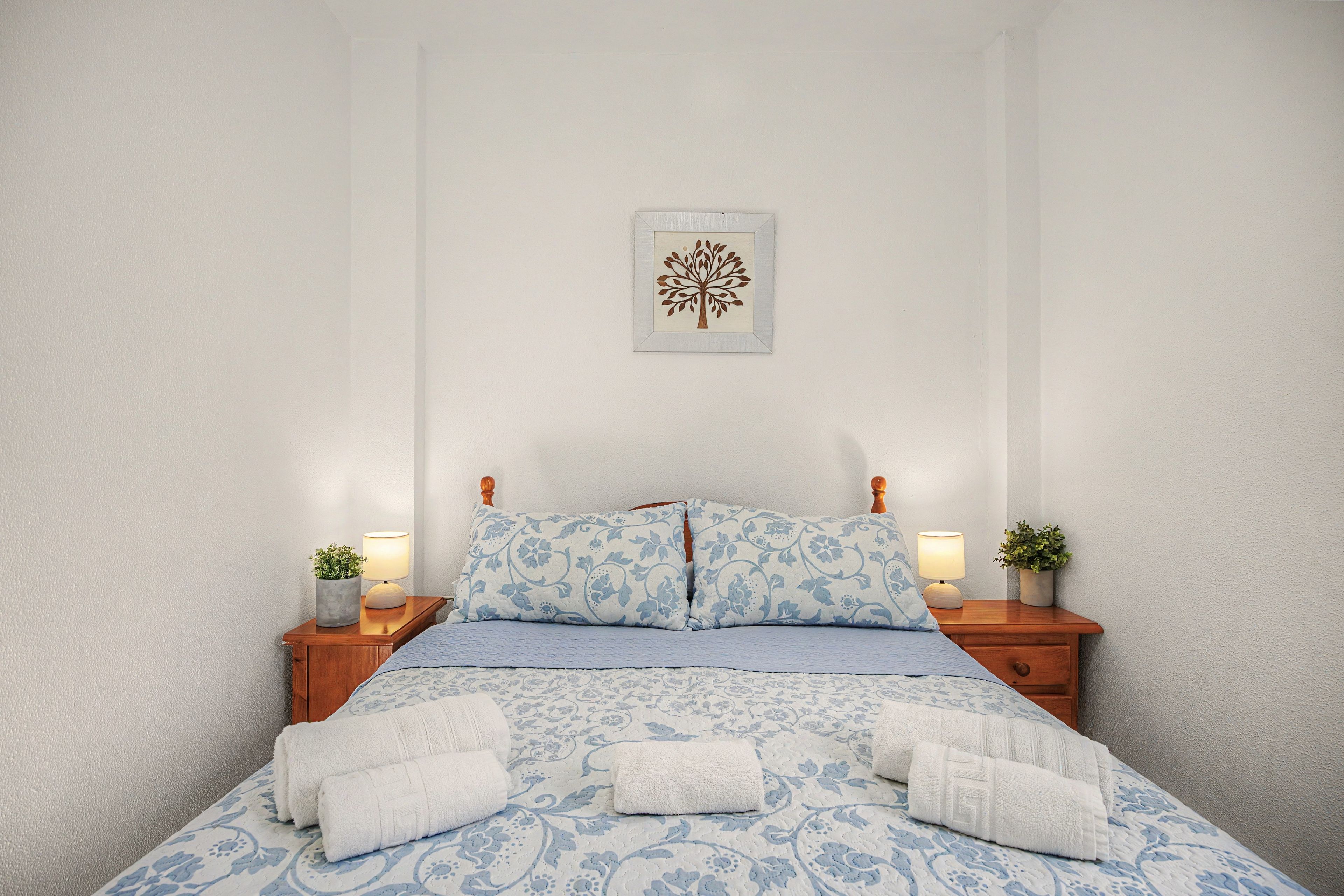 Een net opgemaakt bed met een geborreld sprei, twee kussens en twee nachtkastjes met lampen en plantenpotten.
