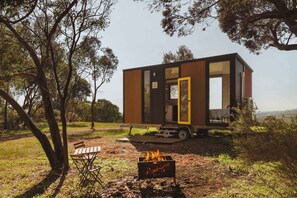 A modern, compact mobile home with a large sliding door, situated in a natural setting with trees and a bench nearby.