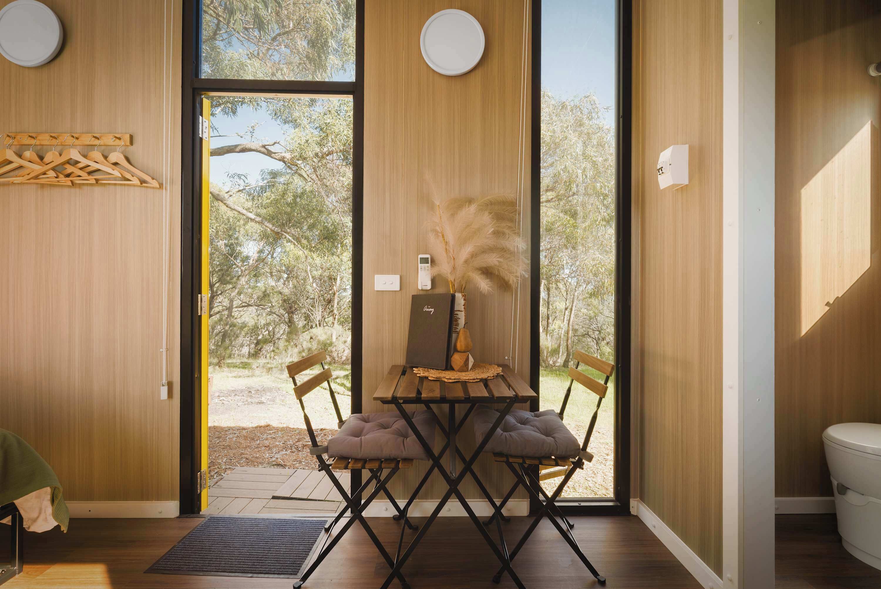 A modern interior with a wooden dining table, two cushioned chairs, a small wooden bench, and a view of the outdoors through large glass doors.