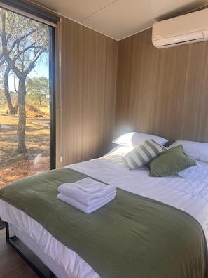 A bedroom with a bed, pillows, a green blanket, a neatly folded towel, and an air conditioning unit. - Tallarook Hilltop Tiny House 5 by Tiny Away (Tallarook)