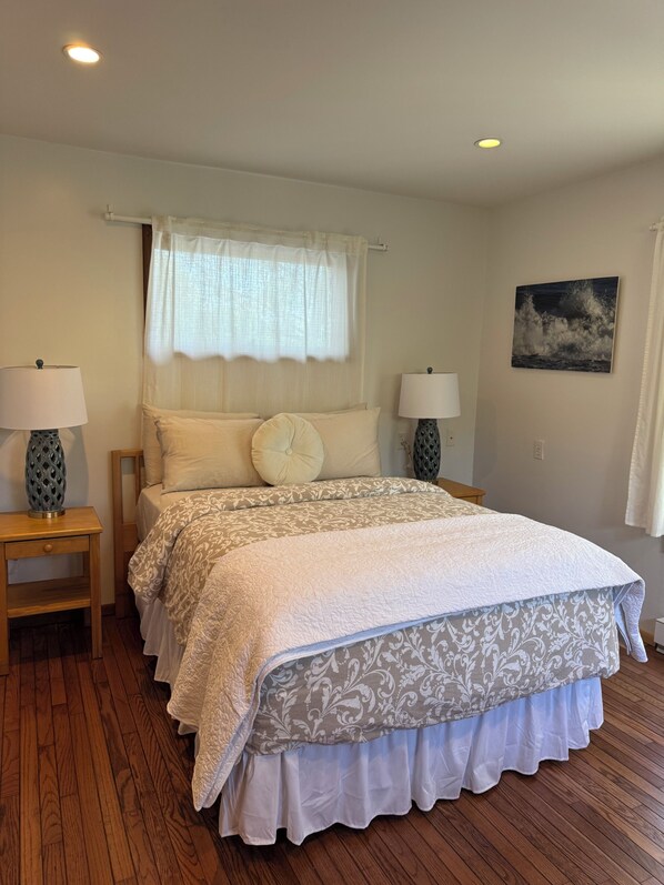 A bedroom with a bed, two bedside tables, a window with curtains, a wall-mounted painting, and a ceiling light.