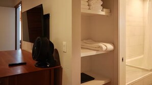 A hotel room with a wooden desk, a black fan, a mirror, a shelf with folded towels, and a shower area.
