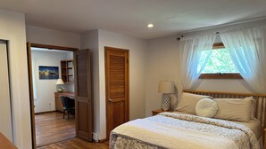 A bedroom with a bed, a nightstand, a lamp, a window with curtains, and a door leading to a hallway.