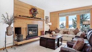 A cozy living room with a fireplace, a sofa, a coffee table, and a large window with a view. - The Buffalo Lodge | Hot Tub • Stunning Mtn Views! (Silverthorne)