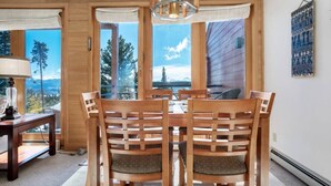 A dining area with wooden chairs and a table, large windows with curtains, and a view of mountains. - The Buffalo Lodge | Hot Tub • Stunning Mtn Views! (Silverthorne)