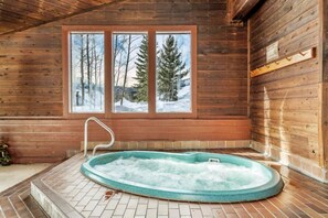 Innenansicht einer Holzhütte mit Whirlpool, einem Fenster mit Blick auf eine verschneite Landschaft und einer Holzbalkendecke.