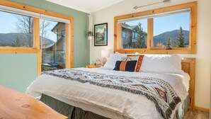 Ein Schlafzimmer mit einem Bett, zwei Fenstern, einem Nachttisch, einer Lampe und Blick auf Berge.