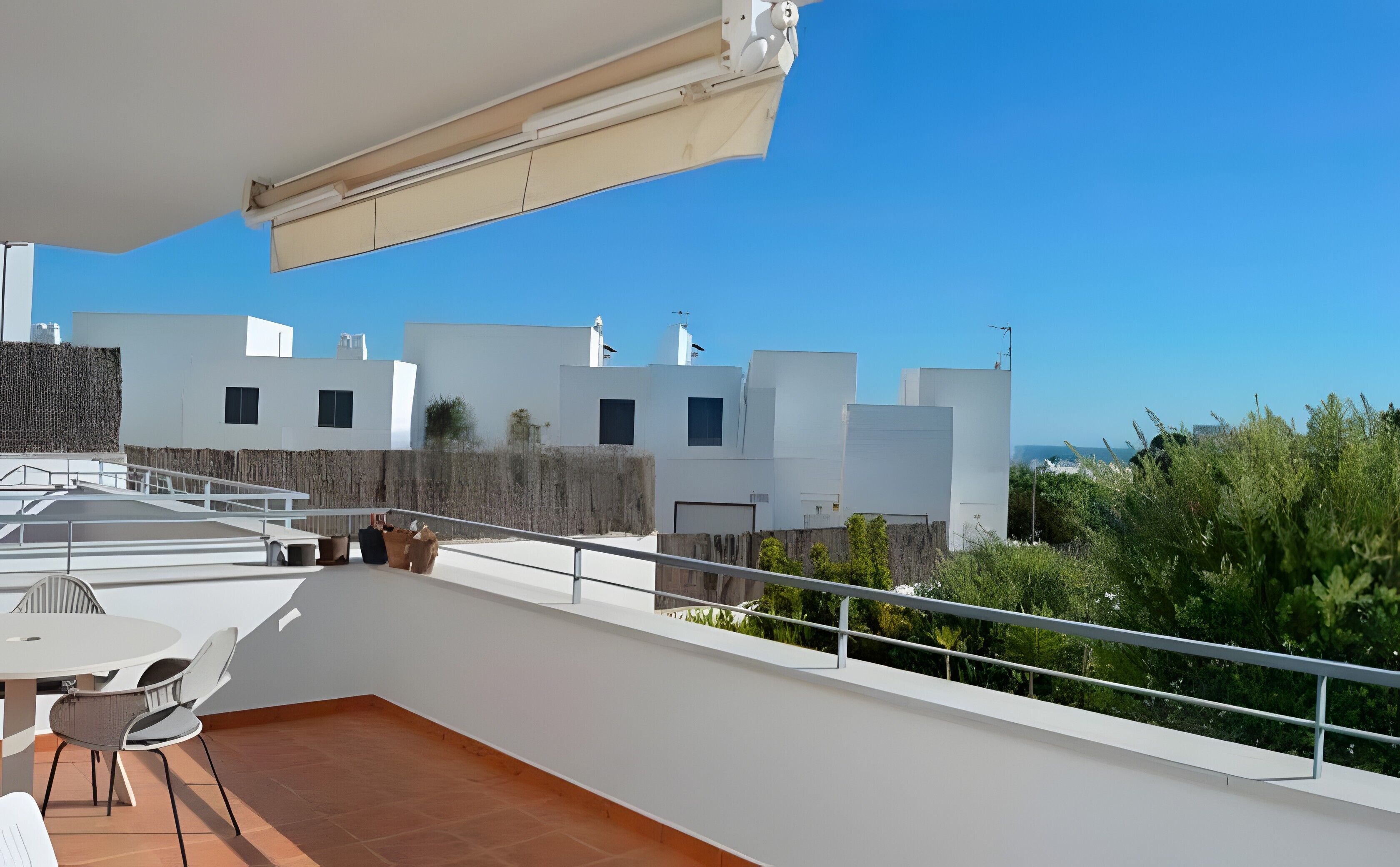 A rooftop terrace with a table and chairs, a railing, and a view of white buildings and greenery.