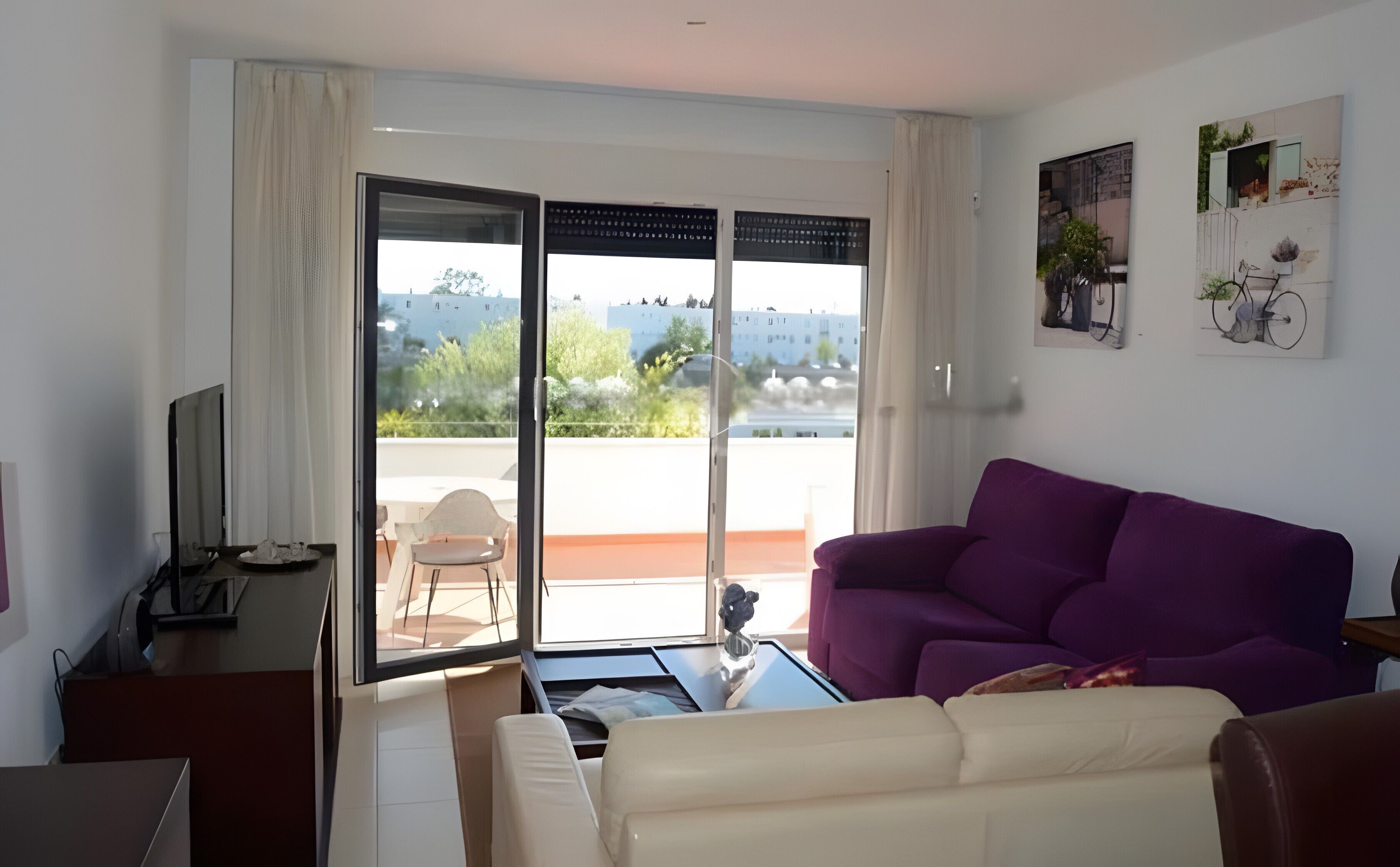 A living room with a purple sofa, a white sofa, a glass table, and a TV.