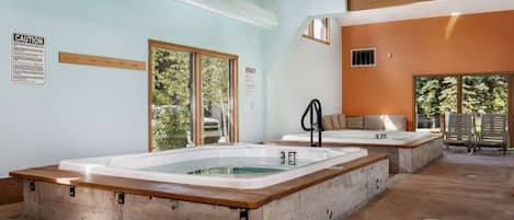 A spa area with two large tubs, a wooden bench, and a view of trees through the windows.