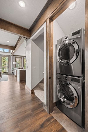 A laundry room with stacked front-loading washers and dryers, wooden beams on the ceiling, and a view of a kitchen and living area through an open door. - Alpine Ambiance | Luxury 4BR • 3 Decks • Hot Tub (Keystone)