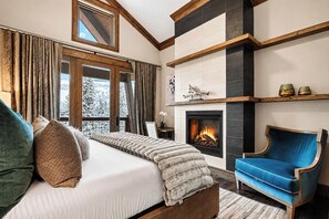 Ein Schlafzimmer mit einem Bett, einem blauen Sessel, einem Kamin und Blick auf schneebedeckte Bäume.