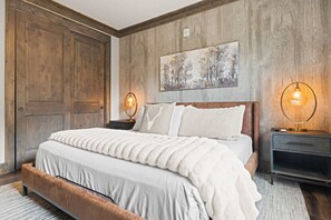 A bedroom with a large bed, wooden walls, a painting, and two bedside lamps. - Alpine Ambiance | Luxury 4BR • 3 Decks • Hot Tub (Keystone)