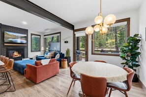 A modern living room with a fireplace, blue sofas, a dining table, and a large window with a view of trees. - All Seasons Retreat | Private Hot Tub | Steam Room (Keystone)