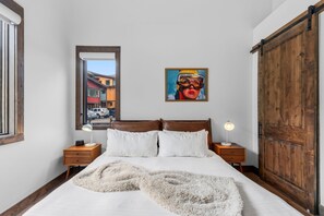 Ein Schlafzimmer mit einem großen Bett, zwei Nachttischen, einem Holzschrank, einem Fenster mit Blick auf Gebäude und einem Bild an der Wand.