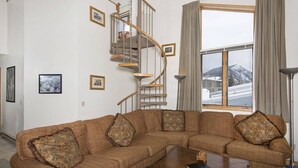 A living room with a curved staircase, a large window with a view, and a sectional sofa with patterned cushions. - Slopeside | Hot Tub • Garage • Pool • Ski Locker! (Keystone)