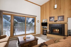 A living room with a fireplace, a television, and a large window with a view of trees. - Slopeside | Hot Tub • Garage • Pool • Ski Locker! (Keystone)