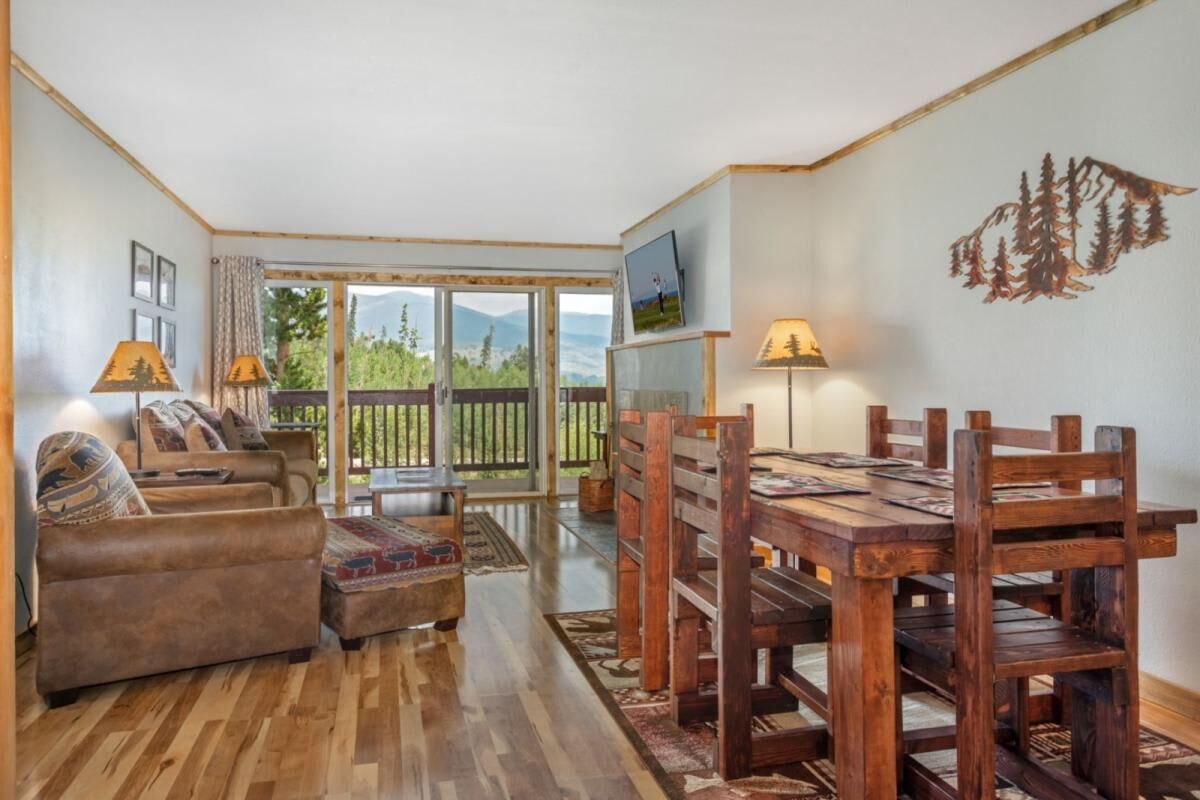 Una habitación con piso de madera, un sofá, una mesa de comedor con sillas, un televisor y vista a las montañas a través de un ventanal.