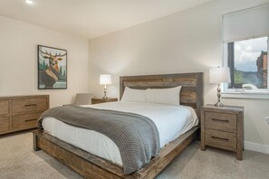 A bedroom with a wooden bed frame, a nightstand with a lamp, a dresser, and a painting of a deer on the wall. - The Sail Loft • Ideal Location • Near Six Resorts! (Dillon)