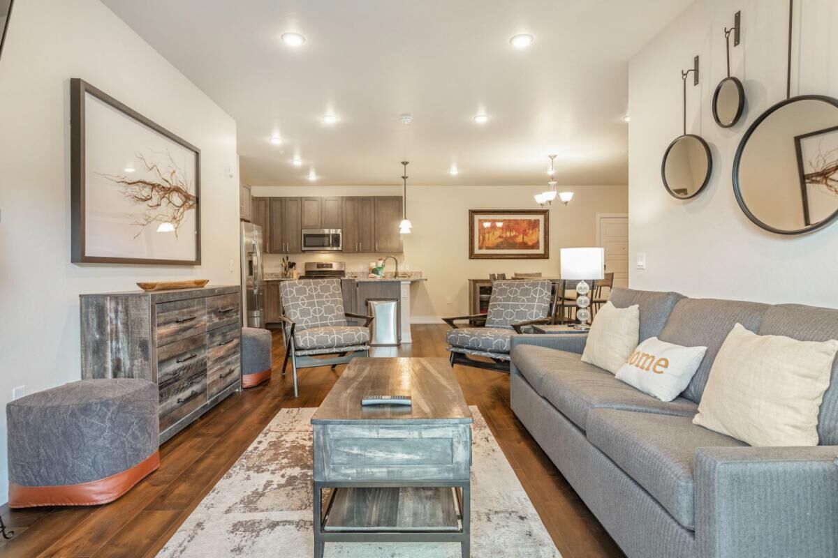 A modern living room with a sofa, ottoman, coffee table, and chairs. There is a kitchen in the background with cabinets and appliances.