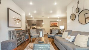 Un salon moderne avec un canapé, un pouf, une table basse et des fauteuils. On aperçoit une cuisine à l’arrière-plan, avec des placards et des appareils électroménagers.