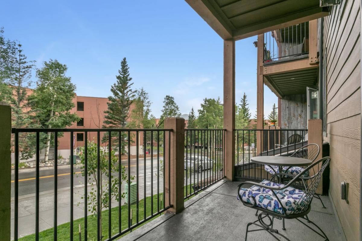 A balcony with a table and chairs, a view of a street and trees, and a building with a balcony.