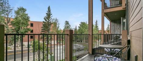 A balcony with a table and chairs, a view of a street and trees, and a building with a balcony.