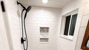 Un bagno piccolo rivestito di piastrelle bianche, con doccia, finestra e lavandino.