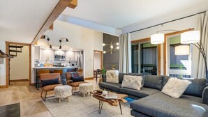 A modern living room with a sofa, coffee table, and chairs, a kitchen in the background, and a large window with curtains. - The Wild Irishman | Hot Tub • Free Shuttle • Pool (Dillon)