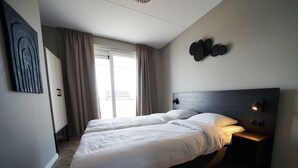 Uma cama de casal com cabeceira, mesa de cabeceira com abajur, guarda-roupa e uma janela grande com cortinas.