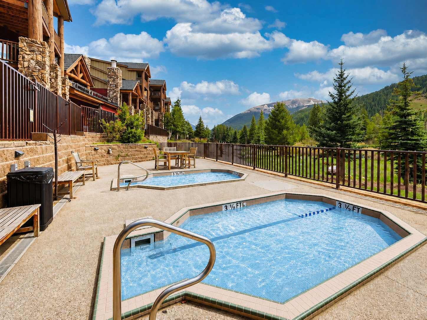 A pool area with two pools, wooden deck seating, and a stone building in the background.