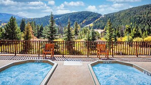 Zona de piscina con dos jacuzzis, una terraza de madera y vistas a una cadena montañosa boscosa.