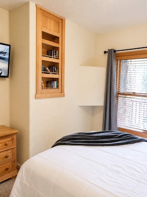 A bedroom with a bed, a wooden bookshelf, a nightstand, a lamp, and a window with curtains. - Slopeside | Hot Tub | Garage | Walk to Lifts! (Keystone)